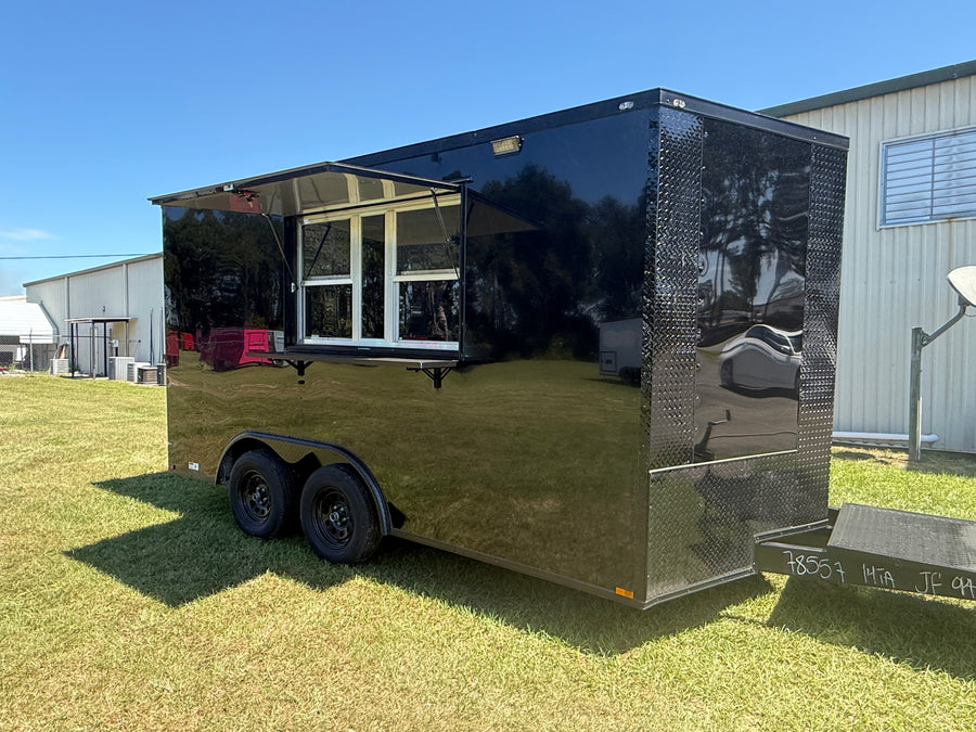 A black food truck trailer parked on grass outdoors, with a serving window extended on the side. The trailer has a glossy reflective black finish, diamond plate side panels, dual wheels, and a hitch at the back.
